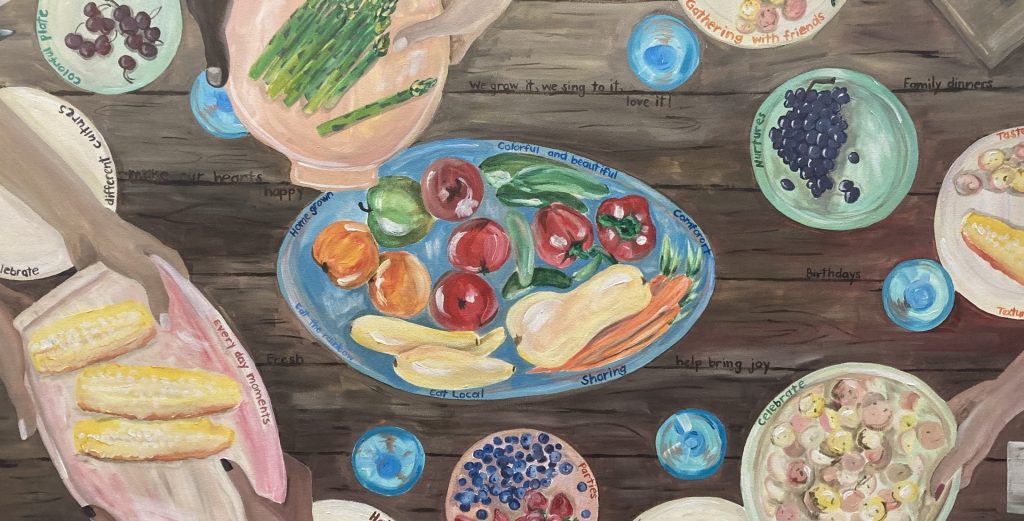 A painting of a meal on a wooden dinner table. At the edges of the painting, hands in many different skin tones reach across the table to share plates of food. In the middle of the table are more plates of colorful fruit and vegetables.